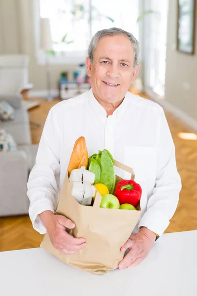 Handsome senior man holding paper bag full of fresh groceries and smiling at home