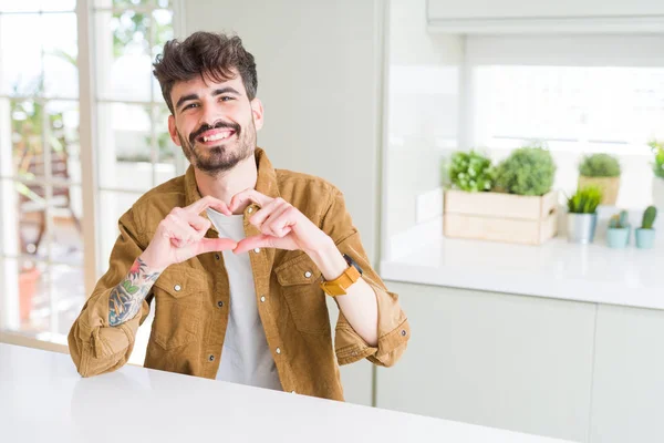 Young man wearing casual jacket sitting on white table smiling in love showing heart symbol and shape with hands. Romantic concept.