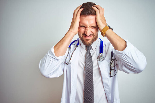 Young handsome doctor man wearing white professional coat over isolated background suffering from headache desperate and stressed because pain and migraine. Hands on head.