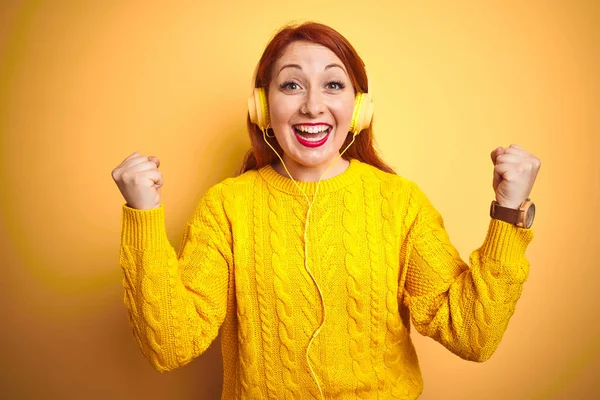 黄色の孤立した背景の上にヘッドフォンを使用して音楽を聴いている若い赤毛の女性は 腕を上げて開いた目で成功に驚き 驚きました 勝者のコンセプト — ストック写真