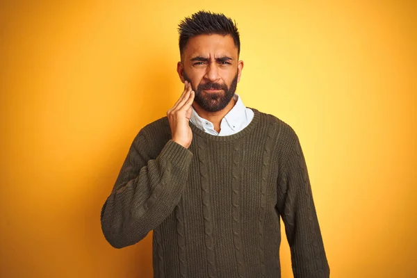 Young indian man wearing green sweater and shirt standing over isolated yellow background touching mouth with hand with painful expression because of toothache or dental illness on teeth. Dentist concept.