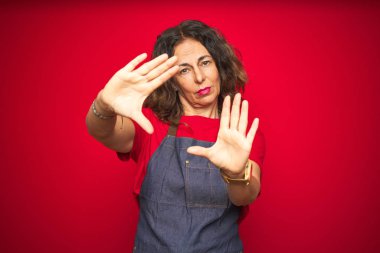 Orta yaş kıdemli kadın el avuç içi ve parmaklar, kamera perspektifi kullanarak çerçeve yapıyor kırmızı izole arka plan üzerinde apron üniforma giyiyor