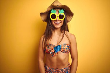 Yüz üzerinde mutlu ve serin bir gülümseme ile izole sarı arka plan üzerinde bikini ve ananas güneş gözlüğü giyen tatil kadın. Şanslı kişi.