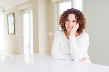 Evde beyaz kazak giyen güzel yaşlı kadın, çapraz kollarla depresyon problemleriyle yorgun ve sıkılmış göründüğünü düşünüyor.