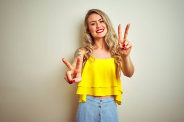 Beyaz izole arka plan üzerinde ayakta sarı t-shirt giyen genç güzel kadın kamera parmaklar zafer işareti yapıyor gösteren görünümlü gülümseyen. İki numara.