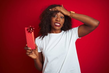 Genç Afrikalı Amerikan kadın tutan Sevgiliye hediye ayakta izole kırmızı arka plan üzerinde el ile vurguladı, utanç ve sürpriz yüz, öfkeli ve sinirli ile şok. Korku ve hata için üzgün.