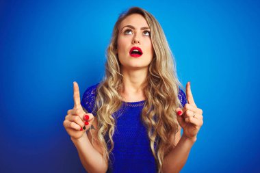 Mavi izole arka plan üzerinde ayakta genç güzel kadın hayret ve görünümlü ve parmaklar ve yükseltilmiş kollar ile işaret şaşırttı.