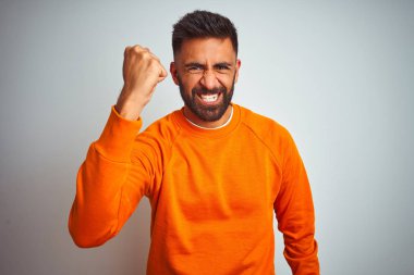 Genç Hint adam üzerinde turuncu kazak giyiyor izole beyaz arka plan öfkeli ve deli yükseltme yumruk sinirli ve öfkeli çığlık sırasında öfke. Öfke ve agresif kavram.