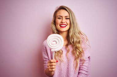 Genç güzel kadın pembe izole arka plan üzerinde mutlu bir yüz ayakta ile tatlı şeker yiyen ve diş gösteren emin bir gülümseme ile gülümseyen