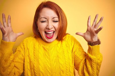 Genç Kızıl saçlı kadın, izole sarı arka plan üzerinde duran kış kazak giyen deli ve silah ile başarı için çılgın kutluyor ve kapalı gözleri heyecanlı çığlık kaldırdı. Kazanan kavramı