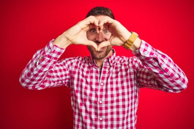 Kırmızı izole arka plan üzerinde genç yakışıklı adam el ve parmaklar işareti ile bakarak gülümseyen kalp şekli yapıyor