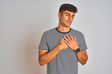 Lacivert çizgili t-shirt giyen genç hintli adam kapalı gözleri ve yüzünde minnettar jest göğüs elleri ile gülümseyerek izole beyaz arka plan üzerinde duran. Sağlık kavramı.