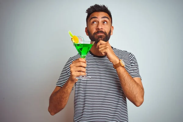 Young indian man drinking cocktail with alcohol standing over isolated white background serious face thinking about question, very confused idea