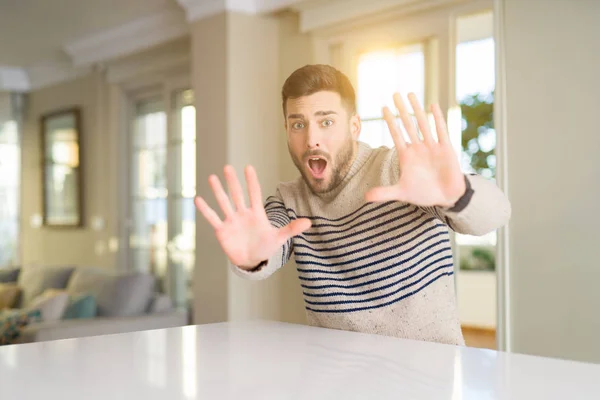 Young handsome man at home afraid and terrified with fear expression stop gesture with hands, shouting in shock. Panic concept.