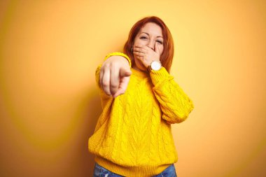 Size gülüyor izole sarı arka plan üzerinde ayakta kış kazak giyen güzel kızıl saçlı kadın, ağız üzerinde el ile kameraya parmak işaret, utanç ifadesi