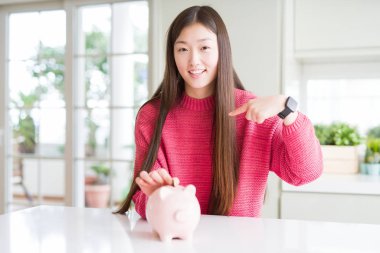 Güzel Asyalı kadın kendisine parmak işaret sürpriz yüz ile Piggy çanta içinde bir sikke koyarak