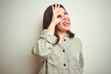 Genç güzel esmer kadın izole arka plan üzerine yeşil gömlek giyen el gülümseyen ok jest yapıyor, göz parmaklarıyla mutlu yüz görünümlü.