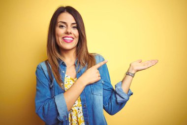 Sarı izole arka plan üzerinde ayakta genç güzel kadın hayran ve el ile sunmak ve parmak ile işaret ederken kameraya gülümsüyor.