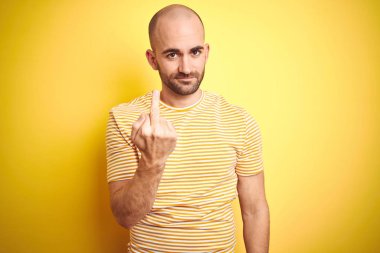 Young bald man with beard wearing casual striped t-shirt over yellow isolated background Showing middle finger, impolite and rude fuck off expression