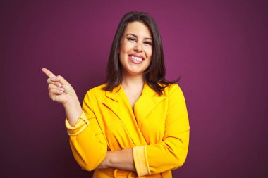 Yüzünde büyük bir gülümseme ile mor izole arka plan üzerinde zarif sarı ceket giyen genç güzel esmer kadın, kamera bakarak yan el ve parmak ile işaret.