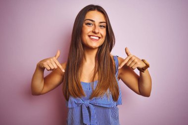 Genç güzel kadın izole pembe arka plan üzerinde ayakta çizgili elbise giymiş yüzünde gülümseme ile emin görünümlü, parmaklarla gurur ve mutlu kendini işaret.