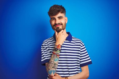 Çizgili polo giyen dövmeli genç adam izole mavi arka plan üzerinde duran kamera güven görünümlü çapraz kollar ve el çene üzerinde kaldırdı gülümseyerek. Olumlu düşünme.