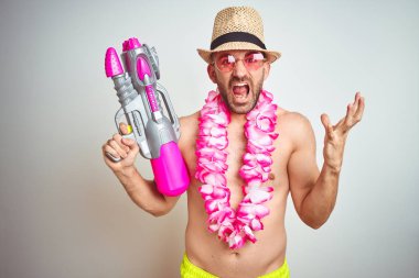 Genç adam yaz şapka giyen ve Hawaii Lei çiçek izole arka plan üzerinde su tabancası tutan çok mutlu ve heyecanlı, kazanan ifade zafer büyük gülümseme ile çığlık kutluyor ve el kaldırdı