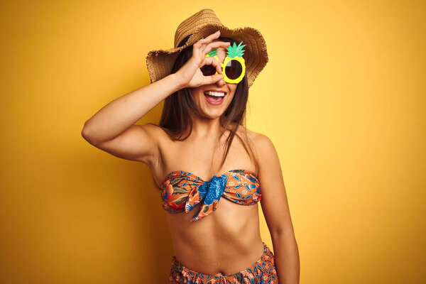 Woman on vacation wearing bikini and pineapple sunglasses over isolated yellow background doing ok gesture with hand smiling, eye looking through fingers with happy face.