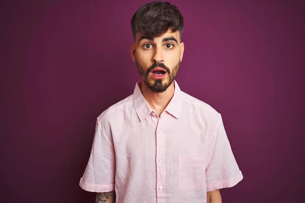 孤立した紫色の背景の上に立つ夏のピンクのシャツを着た入れ墨を持つ若い男は 驚きの表情 恐怖と興奮した顔でショックを受けました — ストック写真
