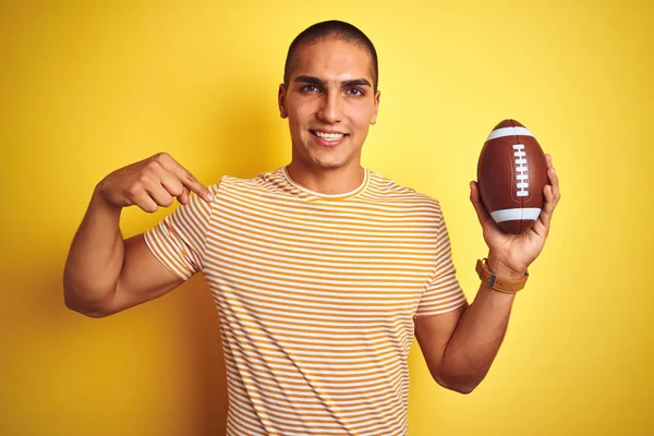 Young rugby player man holding a football ball over yellow isolated background with surprise face pointing finger to himself