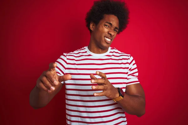 Young african american man with afro hair wearing striped t-shirt over isolated red background disgusted expression, displeased and fearful doing disgust face because aversion reaction. With hands raised. Annoying concept.