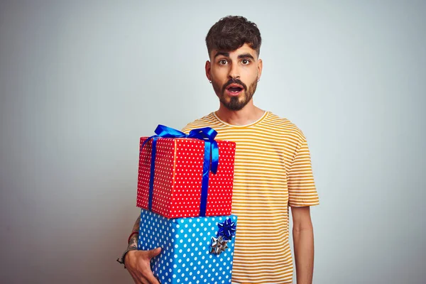 Young man with tattoo holding birthday gifts standing over isolated white background scared in shock with a surprise face, afraid and excited with fear expression