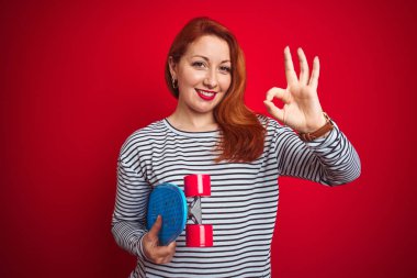 Genç kızıl saçlı öğrenci kadın parmakları ile ok işareti yapıyor kırmızı izole arka plan üzerinde paten tutan, mükemmel sembol