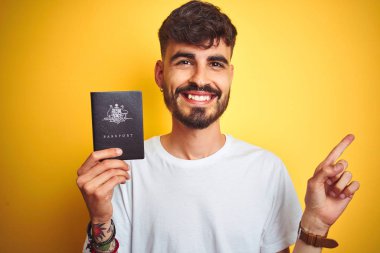 Izole sarı arka plan üzerinde Avustralya Avustralya pasaportu giyen dövme ile genç adam çok mutlu yan el ve parmak ile işaret