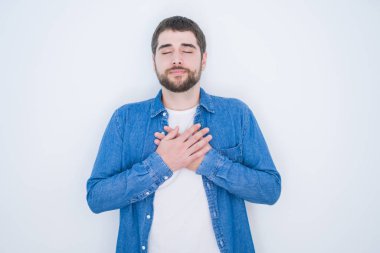 Beyaz izole arka plan üzerine kot ceket giyen genç yakışıklı İspanyol adam kapalı gözleri ve yüz minnettar jest ile göğsüne eller ile gülümsüyor. Sağlık konsepti.