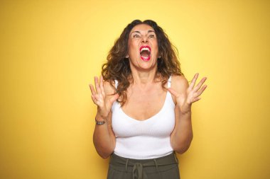 Sarı izole arka plan üzerinde ayakta kıvırcık saçlı orta yaş kıdemli kadın çılgın ve deli çığlık ve agresif ifade ve silah ile bağırdı kaldırdı. Hayal kırıklığı kavramı.