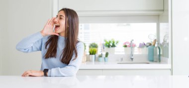 Evde beyaz masada oturan güzel genç kadının geniş açı resim bağırıyor ve ağzına el ile yan gürültülü çığlık. İletişim konsepti.