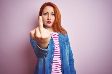 Beautiful redhead woman wearing denim shirt and striped t-shirt over isolated pink background Showing middle finger, impolite and rude fuck off expression