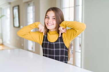 Güzel genç kız çocuk masada oturan yüzünde gülümseme ile kendine güvenen görünümlü, parmaklarıyla gurur ve mutlu kendini işaret.