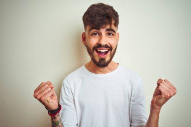 Dövme giyen genç adam izole beyaz arka plan üzerinde duran t-shirt kutlayan şaşırmış ve kollarını kaldırdı ve açık gözleri ile başarı için şaşırttı. Kazanan kavramı.