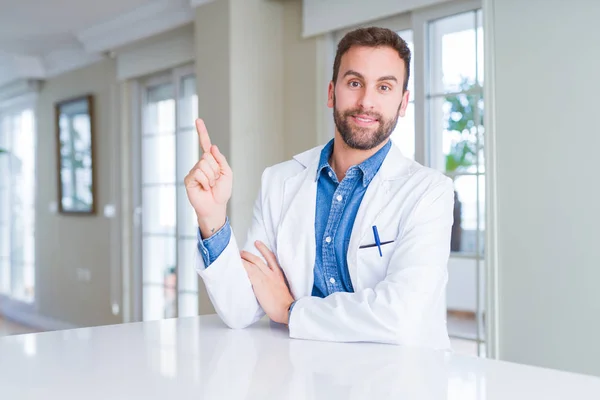 英俊的医生男子穿着医疗外套在诊所与一个大大的微笑在脸上 指着手和手指的一边看着相机 — 图库照片