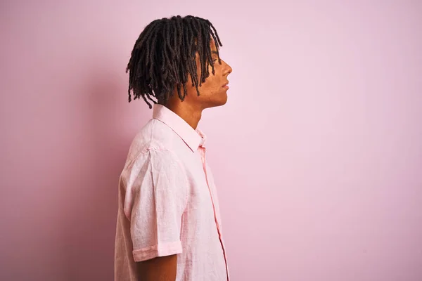 横を見て孤立したピンクの背景の上に立ってエレガントなシャツを着てドレッドロックを持つアフロアメリカ人男性は 自信のある笑顔で自然な顔でプロフィールポーズをリラックス — ストック写真