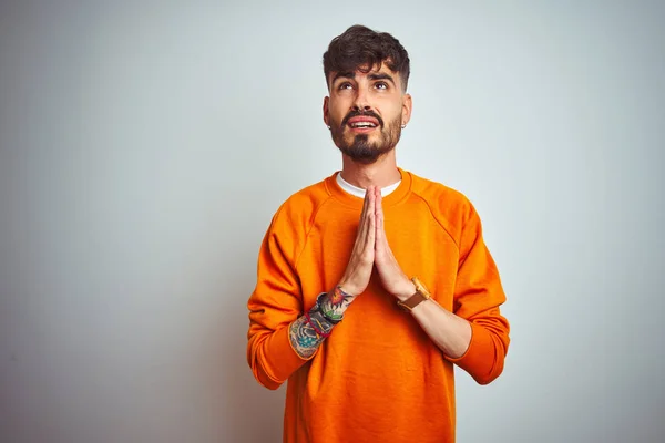 Young man with tattoo wearing orange sweater standing over isolated white background begging and praying with hands together with hope expression on face very emotional and worried. Asking for forgiveness. Religion concept.