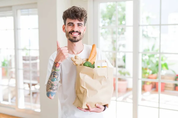 Young man holding paper bag of fresh groceries from the supermarket happy with big smile doing ok sign, thumb up with fingers, excellent sign