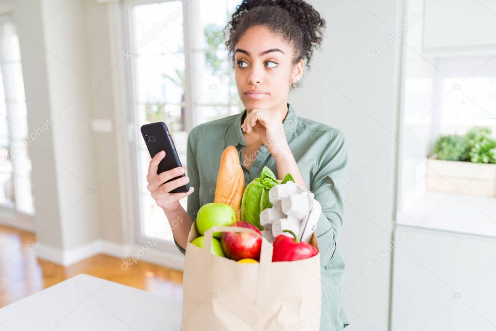 Young african american girl holding paper bag of groceries and using smartphone serious face thinking about question, very confused idea