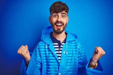 İzole mavi arka plan üzerinde duran yağmurluk giyen genç yakışıklı adam kollarını kaldırdı ve gözleri açık başarı için sürpriz ve şaşırttı. Kazanan kavramı.
