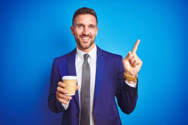Mavi izole arka plan üzerinde bir kağıt bardak üzerinde bir kahve içme genç yakışıklı iş adam çok mutlu yan el ve parmak ile işaret