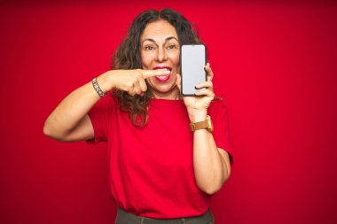 Kırmızı izole arka plan üzerinde akıllı telefonun ekranını gösteren orta yaş kıdemli kadın çok mutlu el ve parmak ile işaret