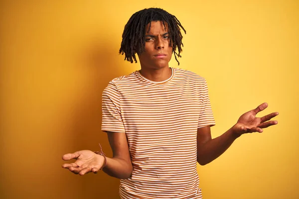 Dreadlocks ile Afro adam izole sarı arka plan clueless üzerine çizgili t-shirt ayakta giyen ve açık kollar ile karıştı, hiçbir fikir kavramı.