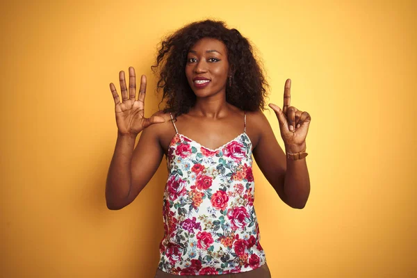 孤立した黄色の背景の上に花柄の夏のTシャツを着たアフリカ系アメリカ人女性は 自信を持って幸せに微笑みながら 指番号7を示し 指を上げる — ストック写真